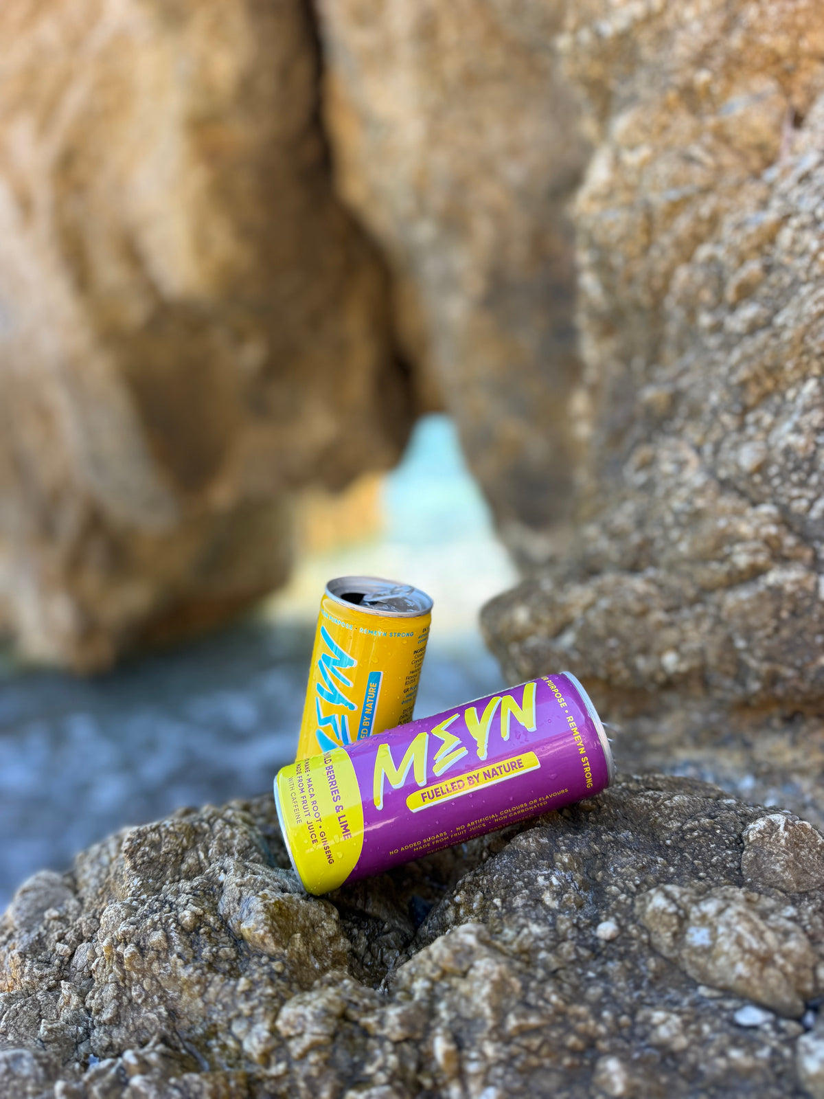 Two Meyn energy drink cans, one yellow and one purple, resting on large rocks outdoors.