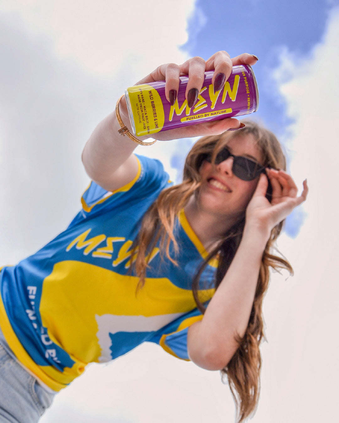 A woman outdoors under a blue sky holds up a purple and yellow can of MEYN energy drink, wearing a blue and yellow sports jersey and sunglasses.